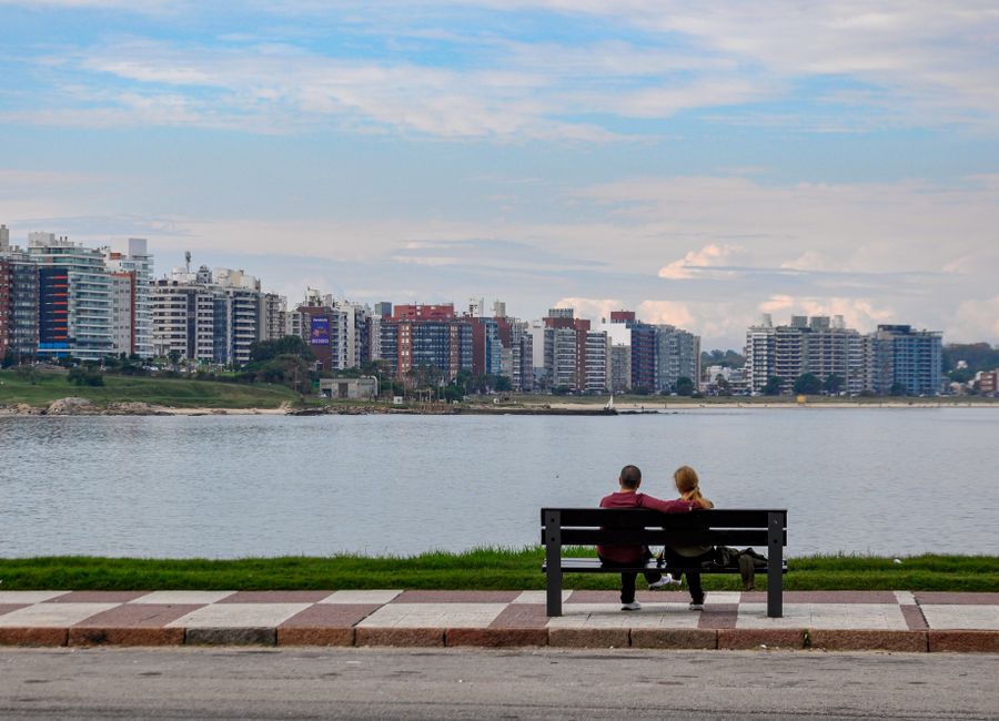 A couple enjoys a serene view of Montevideo's skyline from a waterfront bench.