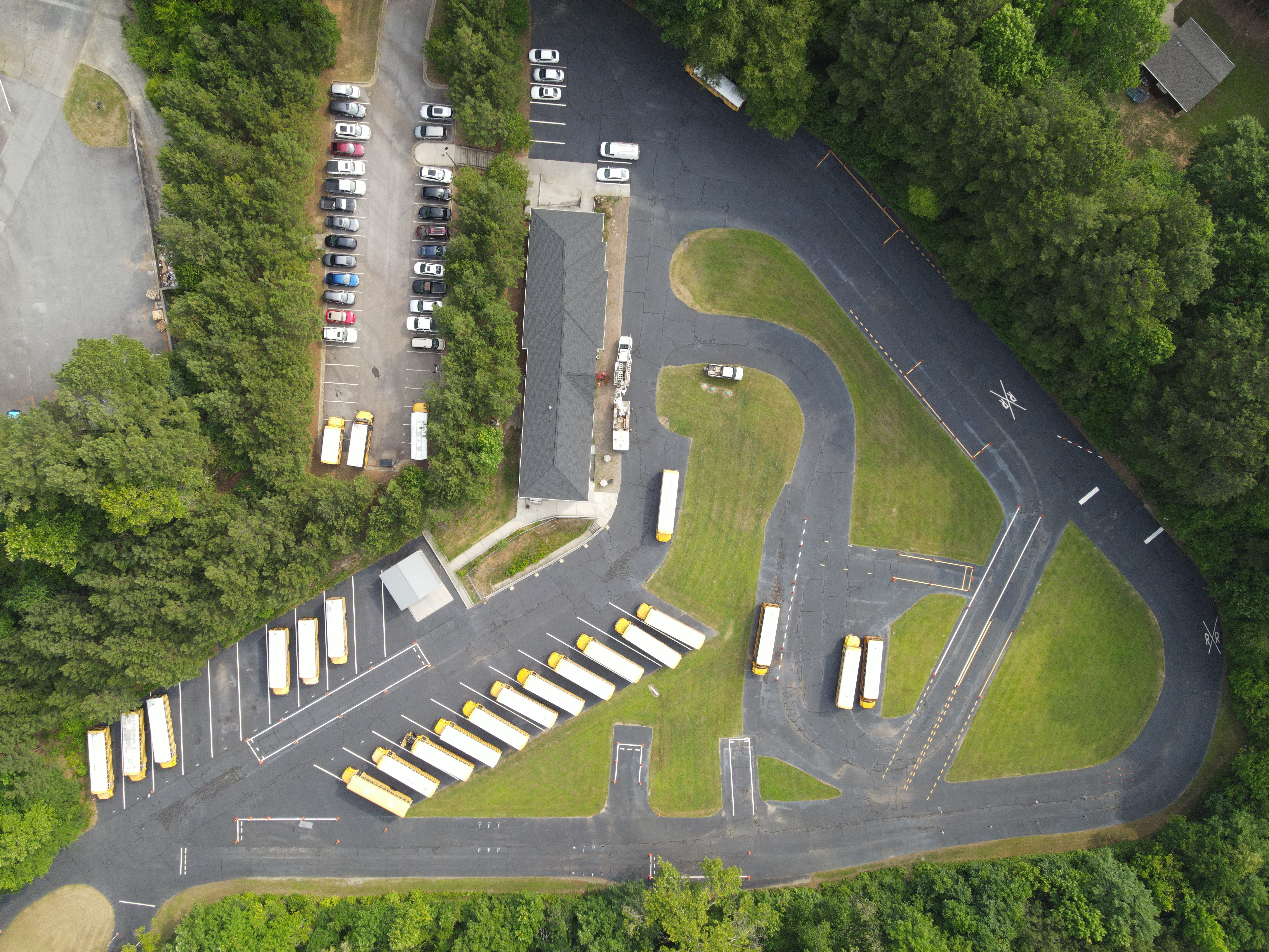 Aerial shot of a school parking lot with multiple yellow school buses and parked cars surrounded by trees.
