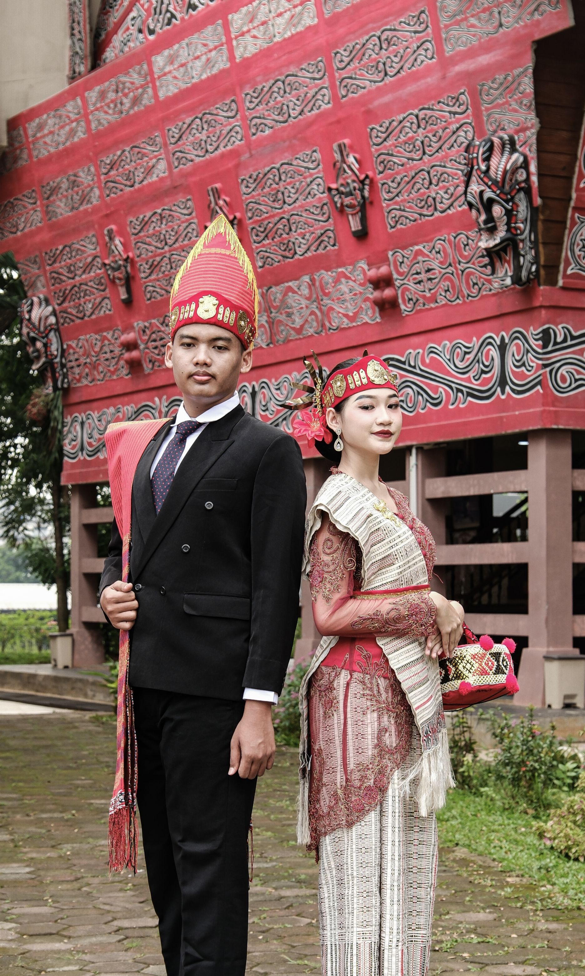 Couple in traditional Batak attire pose by iconic Sumatra landmark. Rich cultural heritage.