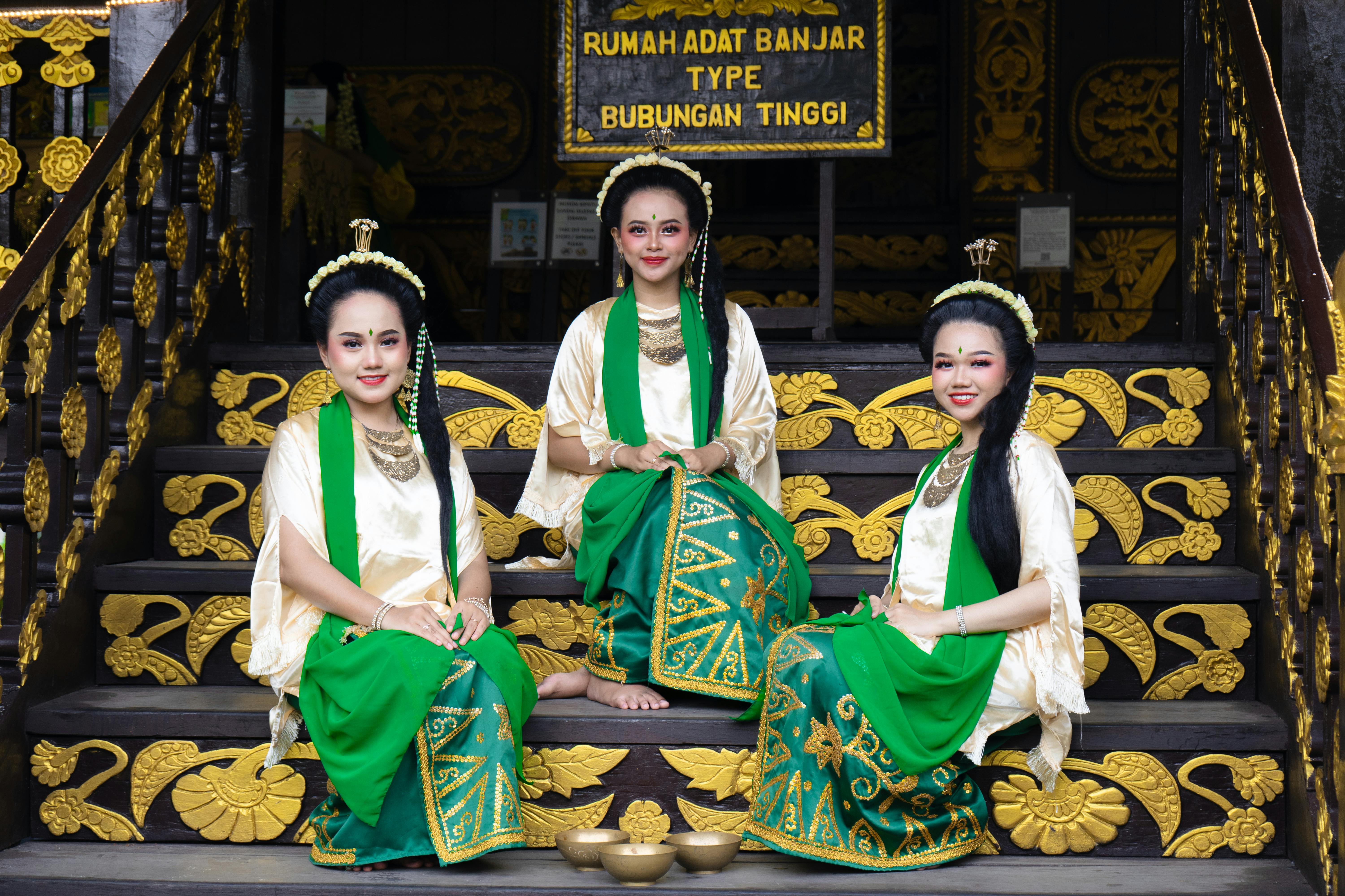 Three women in traditional Banjar attire seated on ornately decorated stairs with intricate designs.