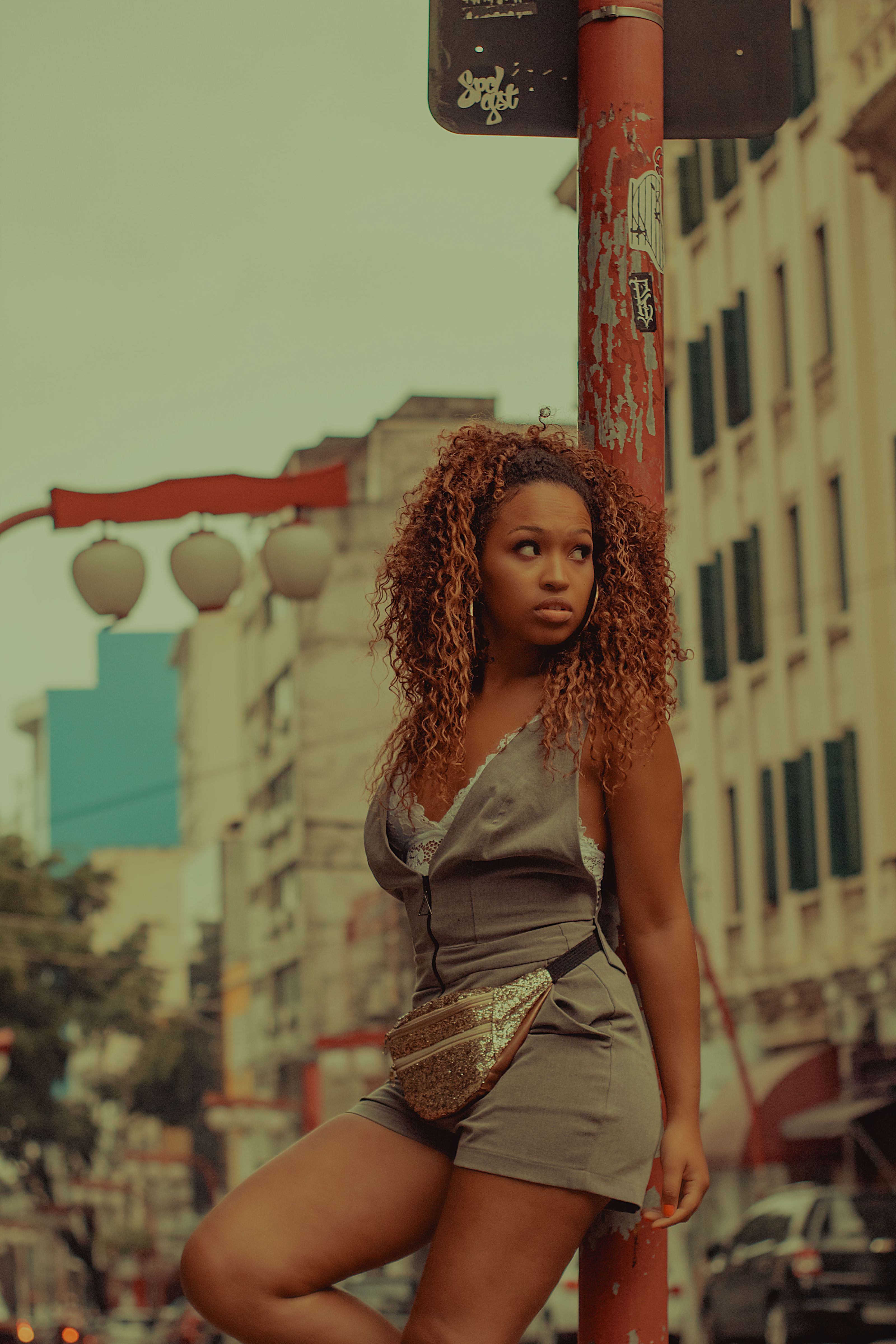 Mulher com cabelo cacheado posando em uma rua da cidade com prédios urbanos ao fundo.