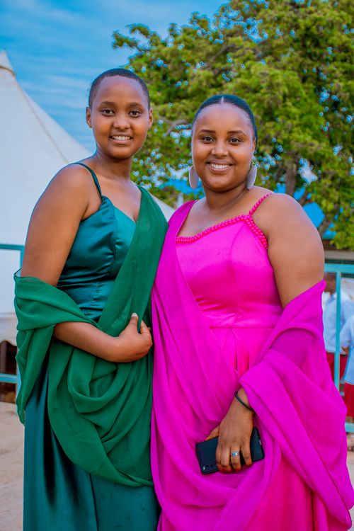 Free Two women in vibrant traditional dresses smiling at an outdoor event. Lush greenery and tents in the background. Stock Photo