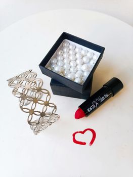 Flat lay of pearls, silver bracelet, and red lipstick on white background.