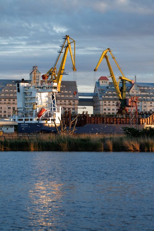 Free A view of cranes and a cargo ship at Kaliningrad port during sunset, reflecting on the water. Stock Photo