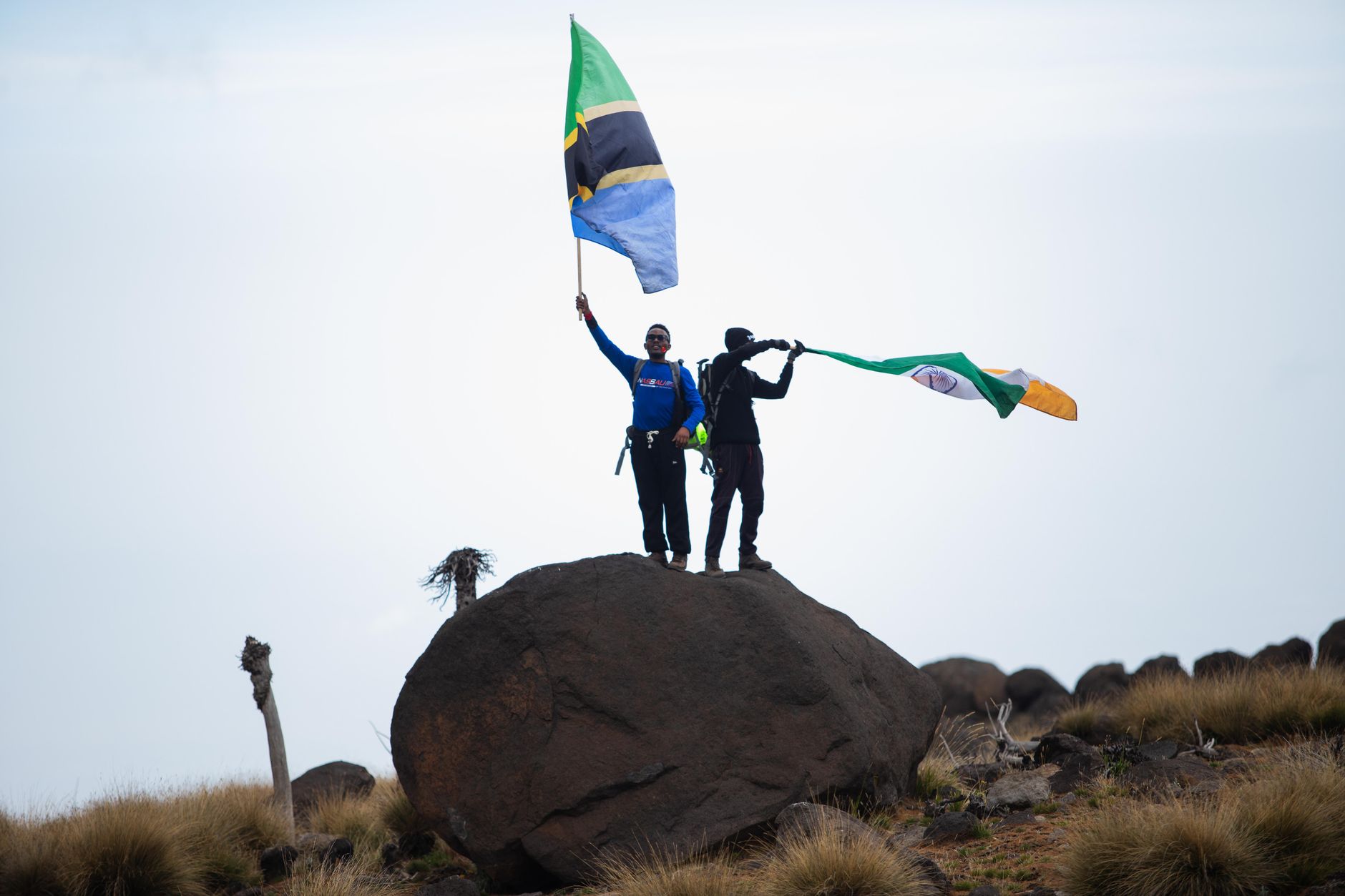 Two adventurers stand victoriously atop Mt. Kilimanjaro, waving flags, symbolizing triumph and international unity.