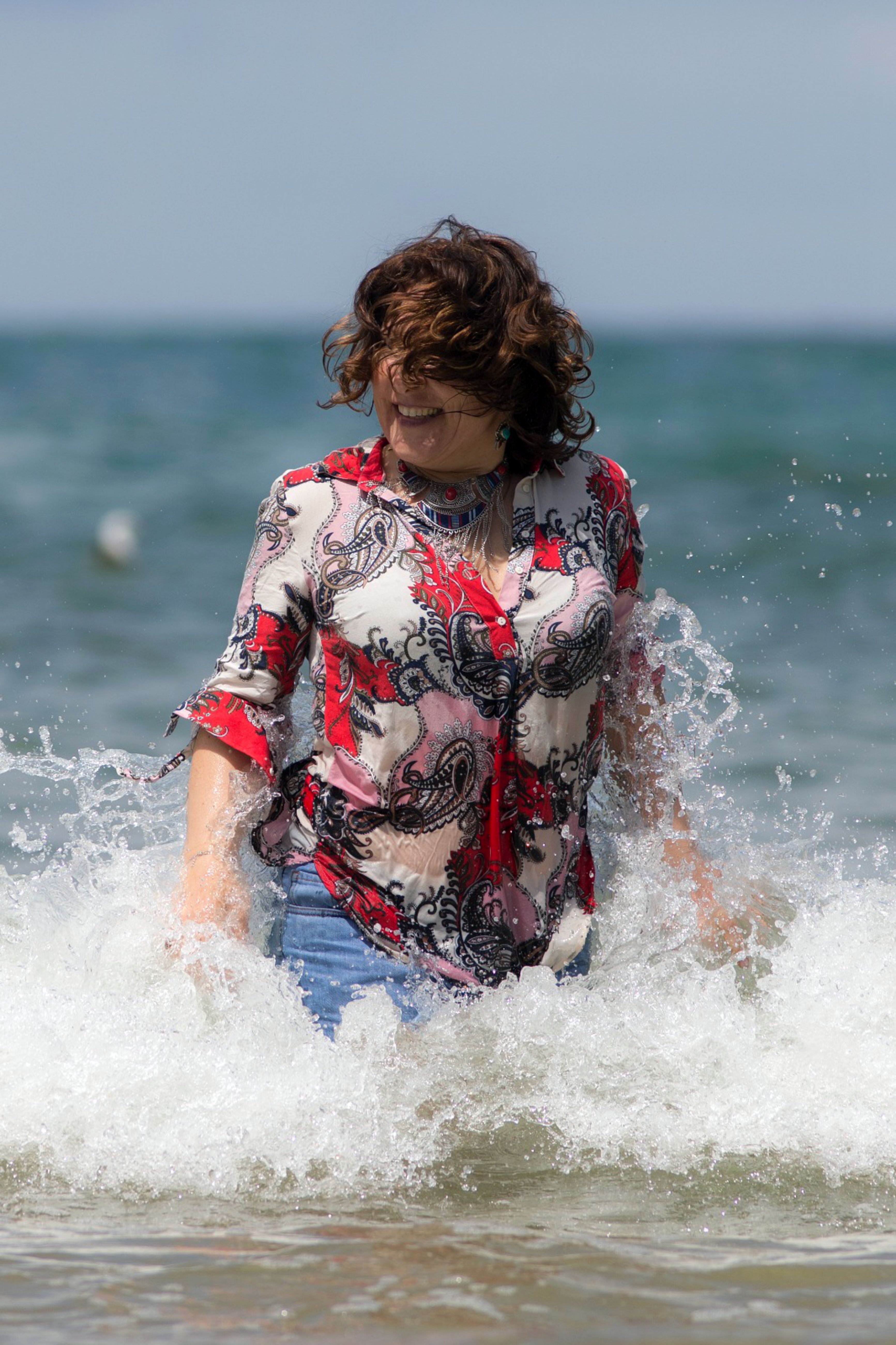 Smiling woman in vibrant attire plays in ocean waves on a sunny Lisbon beach day.