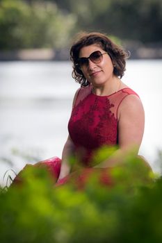 Portrait of a woman in a red dress and sunglasses by a lake on a sunny day.