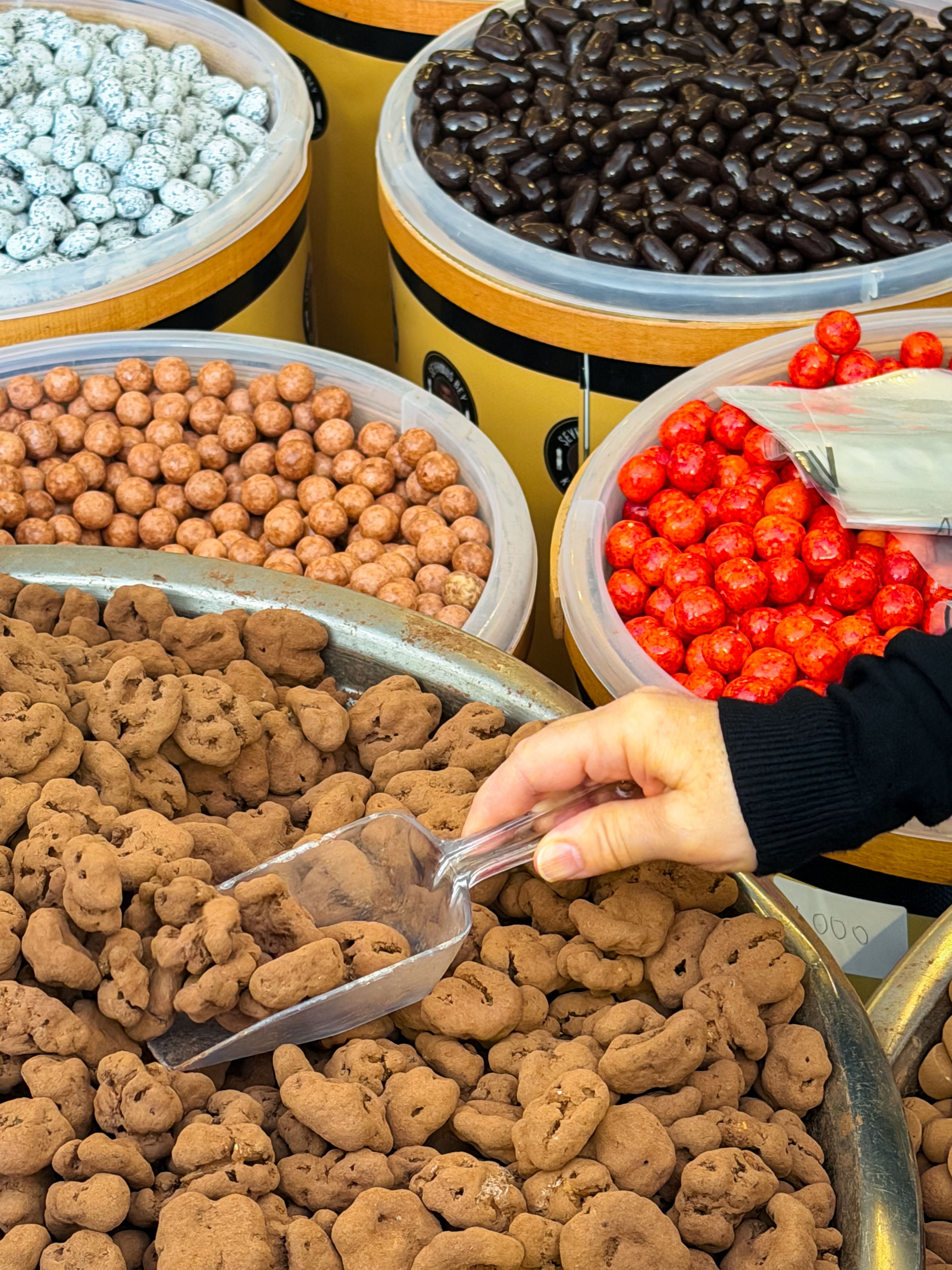 A hand takes a plastic scoop to a bin of cocoa-dusted candies, with tubs of coated sweets behind.