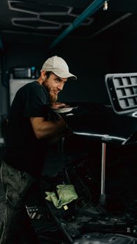 Young male mechanic works on a sleek car hood in a dimly lit garage.