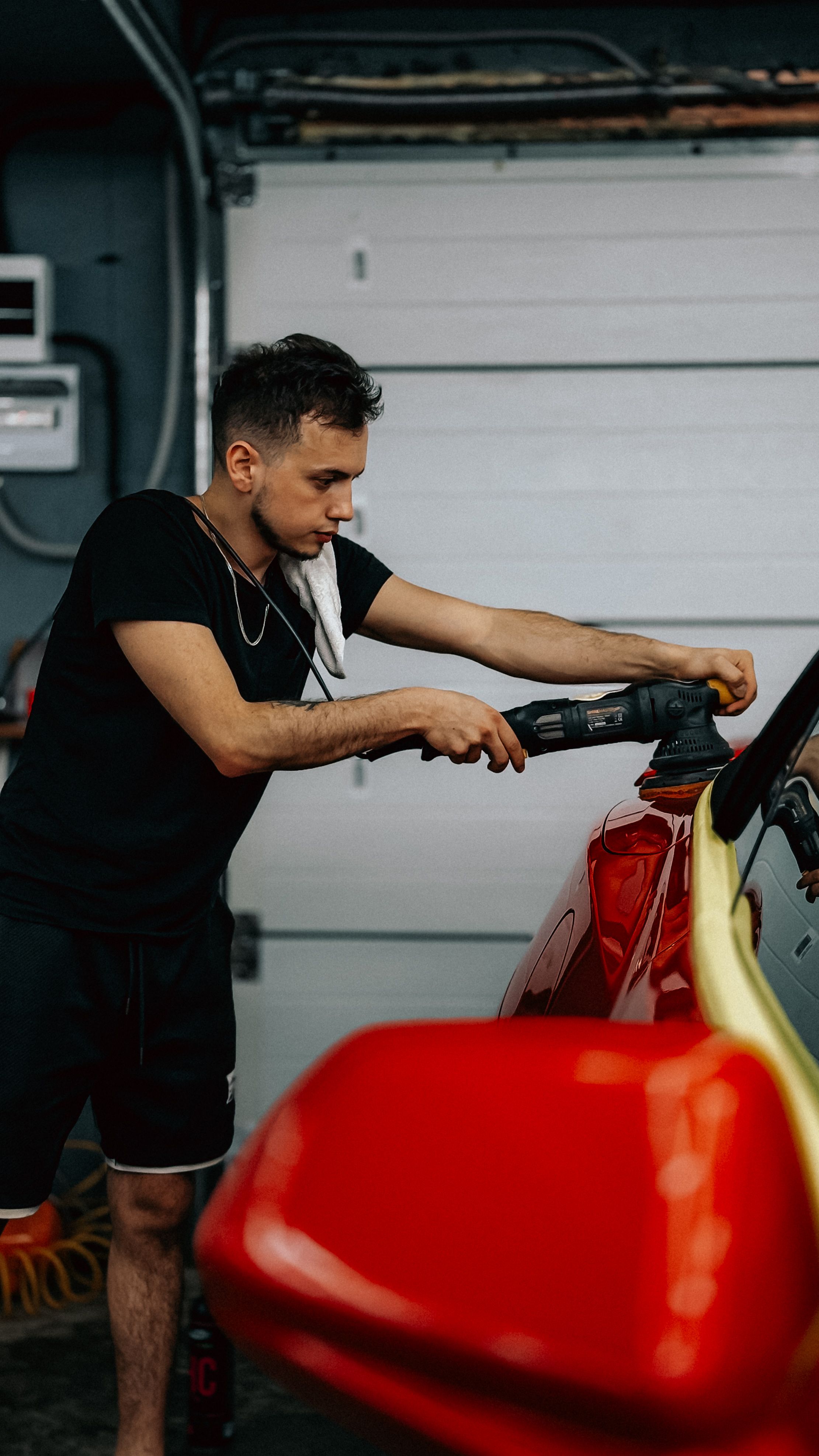 Young adult polishing car detailing red vehicle inside garage workshop.