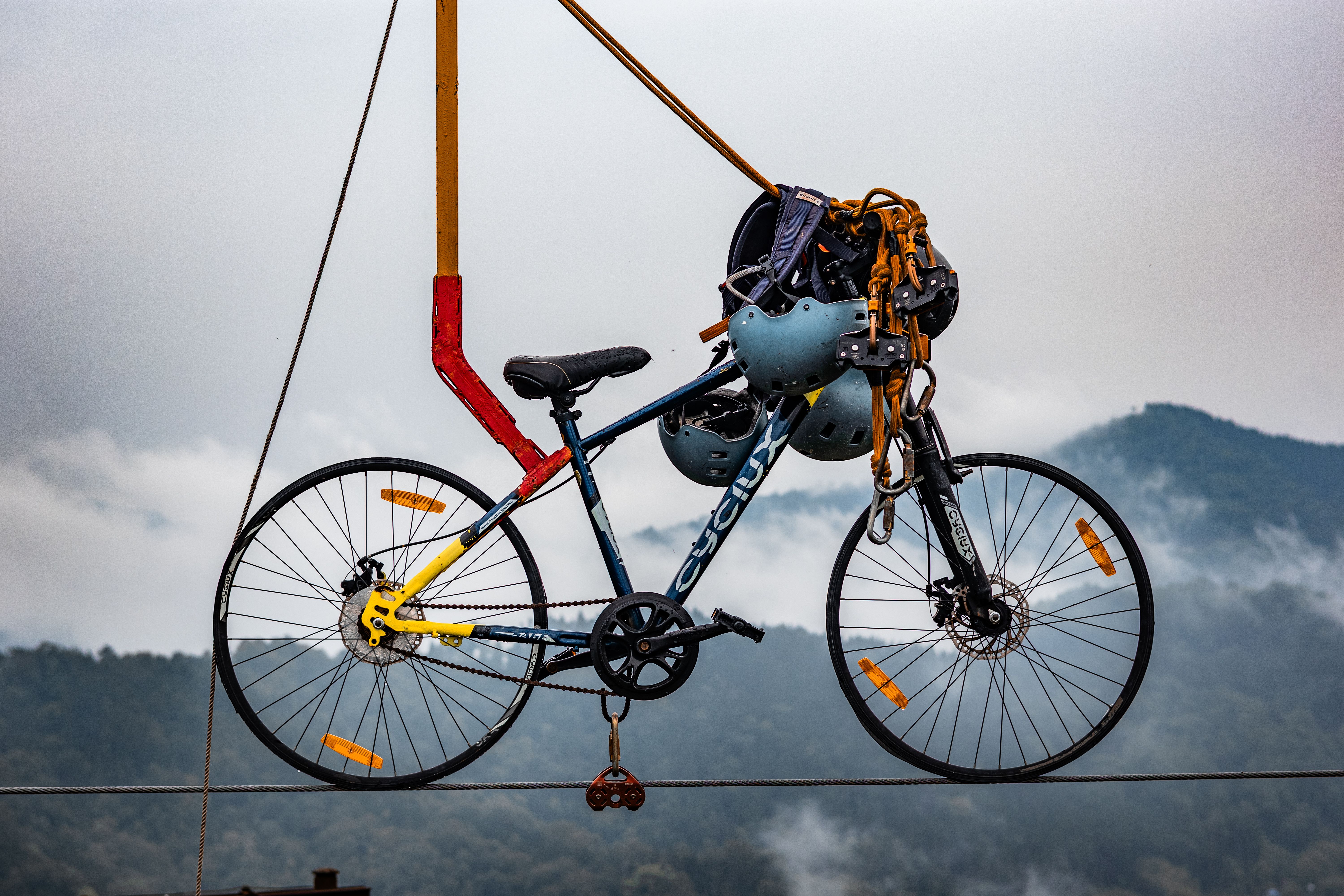 A bicycle suspended on a cable line with helmets against a misty mountainous background.