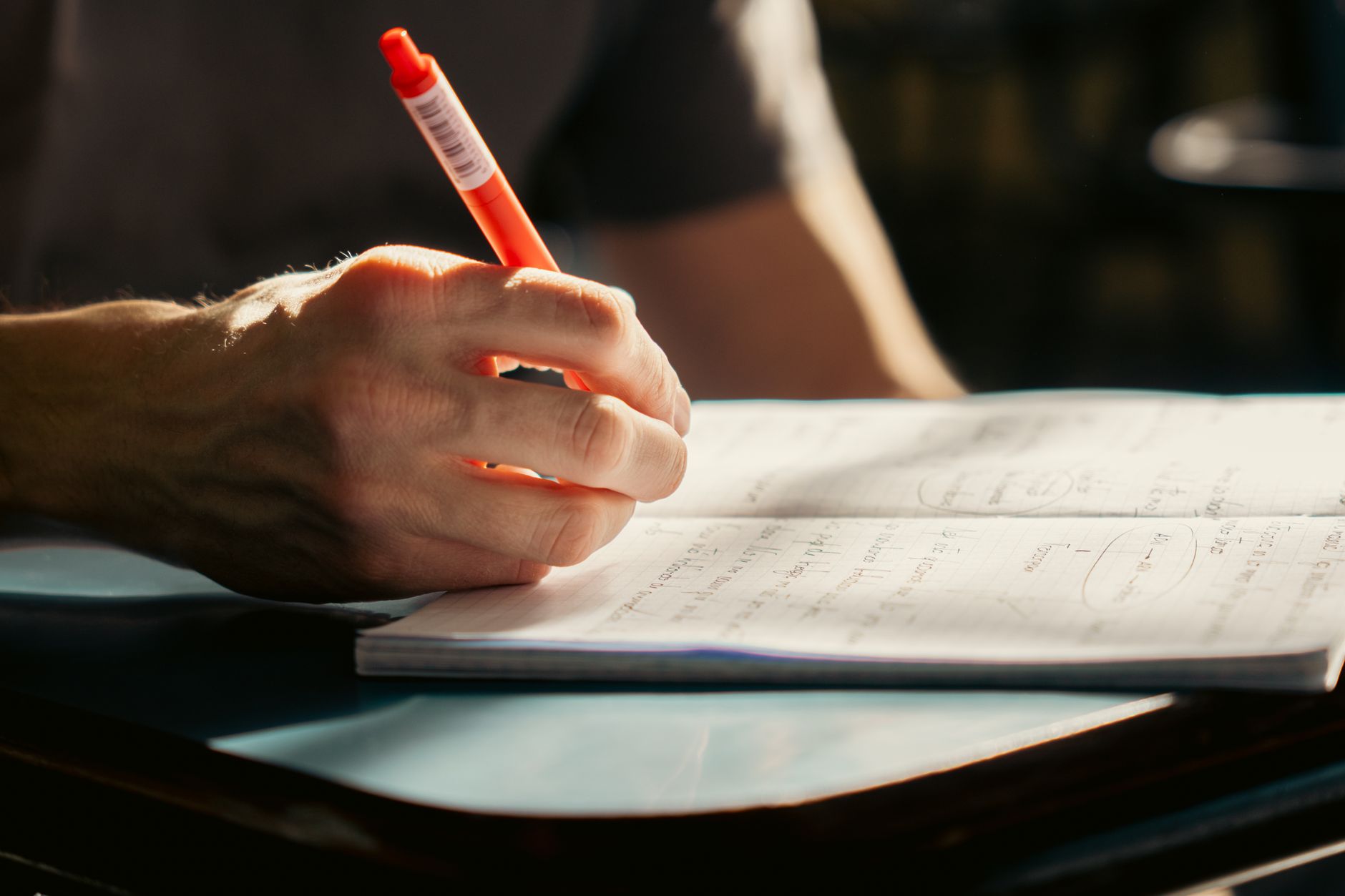 A student writing in a notebook with a pen, in a well-lit classroom environment.