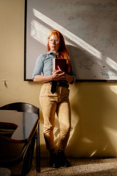 Confident young teacher holding a book in a sunlit classroom with a whiteboard.