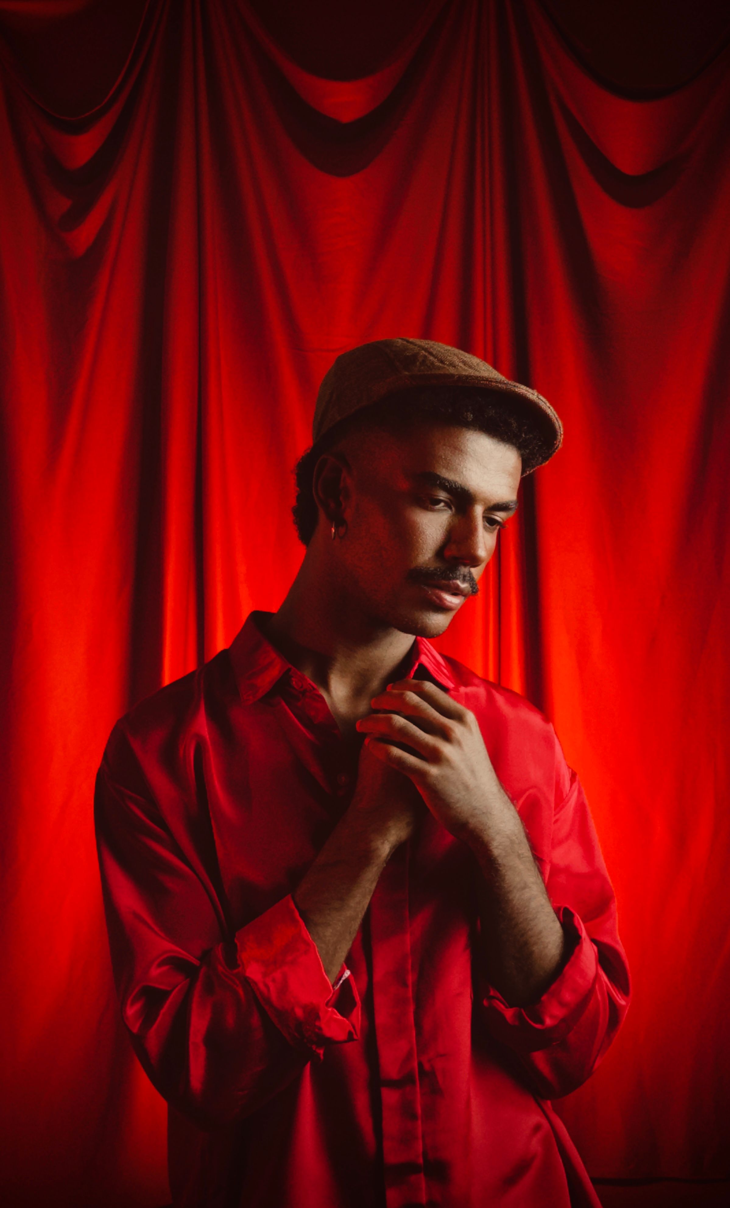 Young man in red shirt standing pensively against red curtain backdrop.