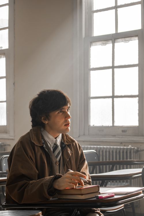 Free A student sits thoughtfully at a desk in a sunlit classroom, holding books and looking upward. Stock Photo