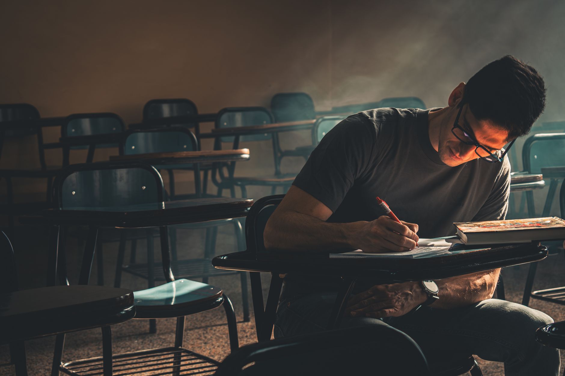 Student focused on writing, studying in a dimly lit empty classroom.