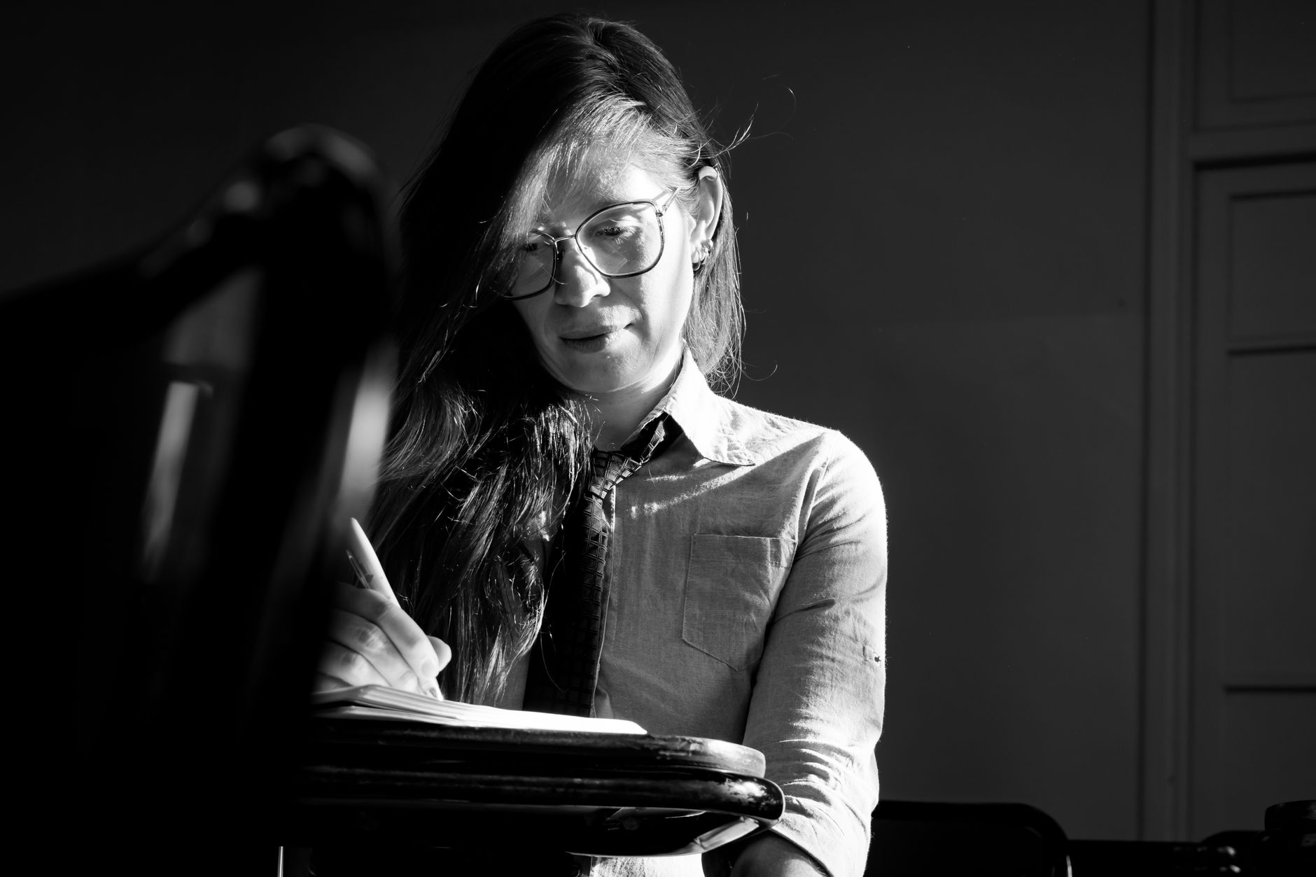 A thoughtful woman writes in a notebook in a shaded classroom, captured in black and white.