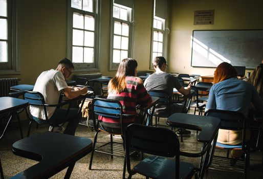 A group of students focused on writing in a sunlit classroom with desks and windows.