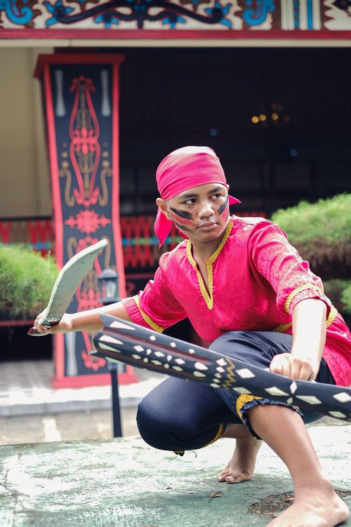 Free Portrait of a young Indonesian man in traditional warrior attire with swords, showcasing cultural heritage. Stock Photo