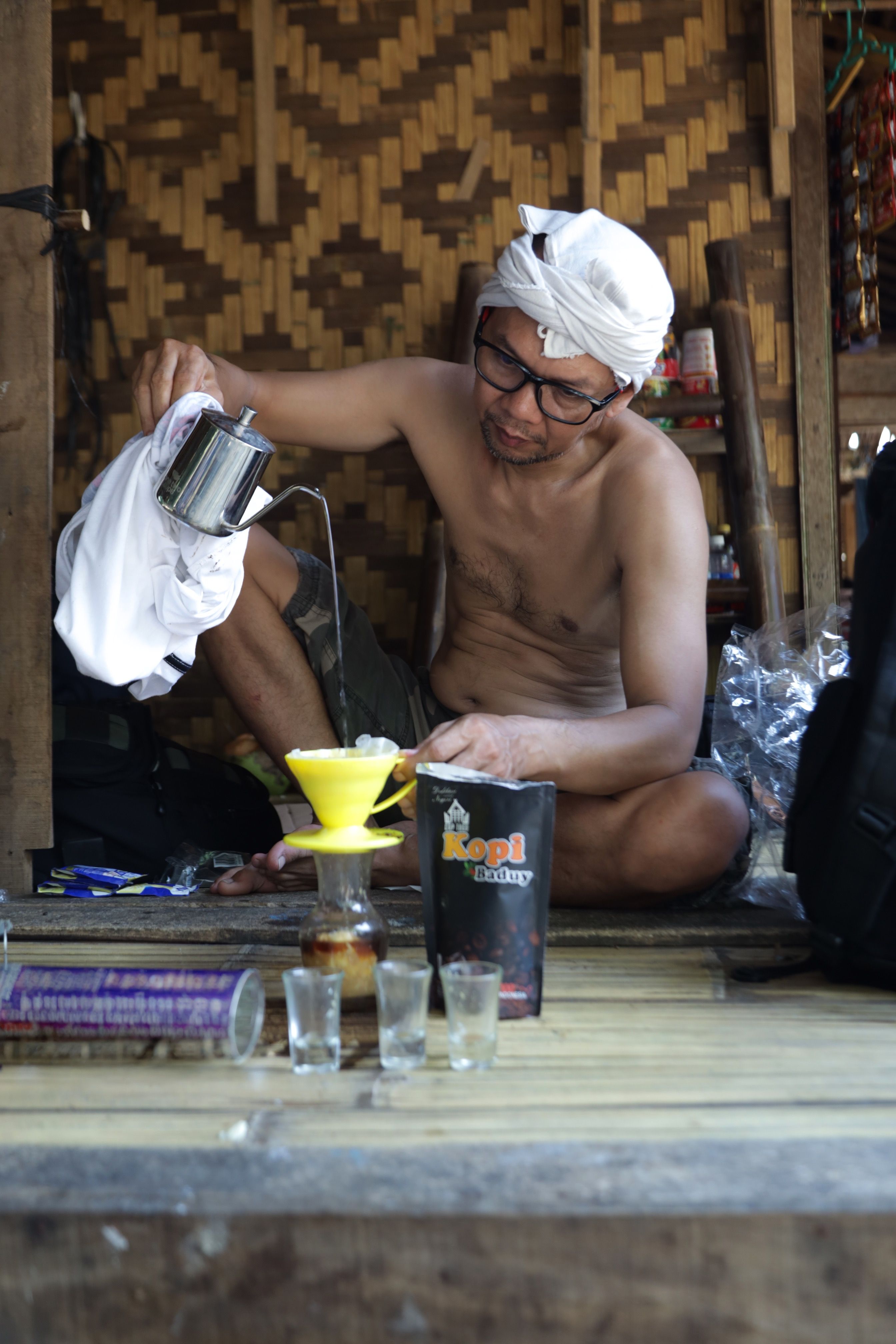 Man brewing coffee in a traditional setting using manual tools and wearing white headscarf.