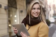 在城市环境中，一位穿着秋装的年轻女子拿着平板电脑微笑