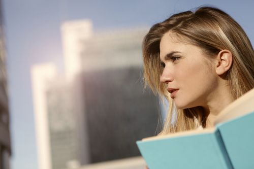 Free Focused young woman reading a book outdoors with city buildings in the background. Stock Photo