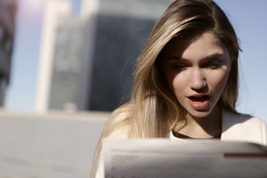 白天，一名金发年轻女子在户外读报时露出了惊讶的表情