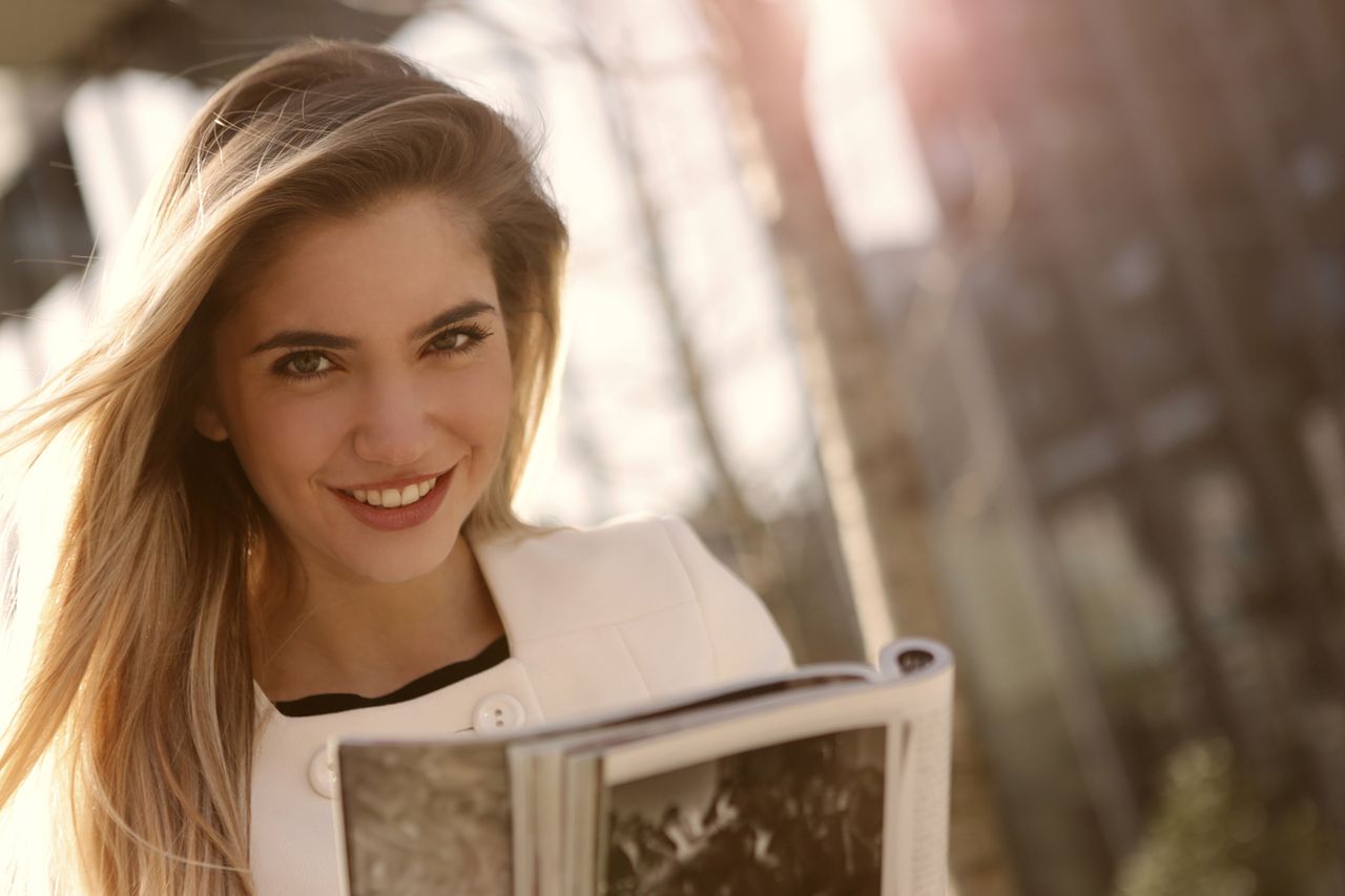 smiling woman with long blond hair reading a magazine outdoors in sunl