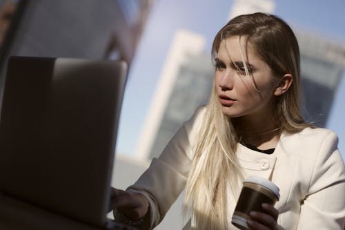 Mujer En Chaqueta Blanca Sosteniendo Una Taza Desechable Mientras Usa La Computadora Portátil