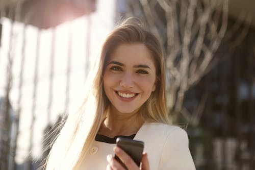 黒のスマートフォンを保持している白いブレザーの笑顔の女性