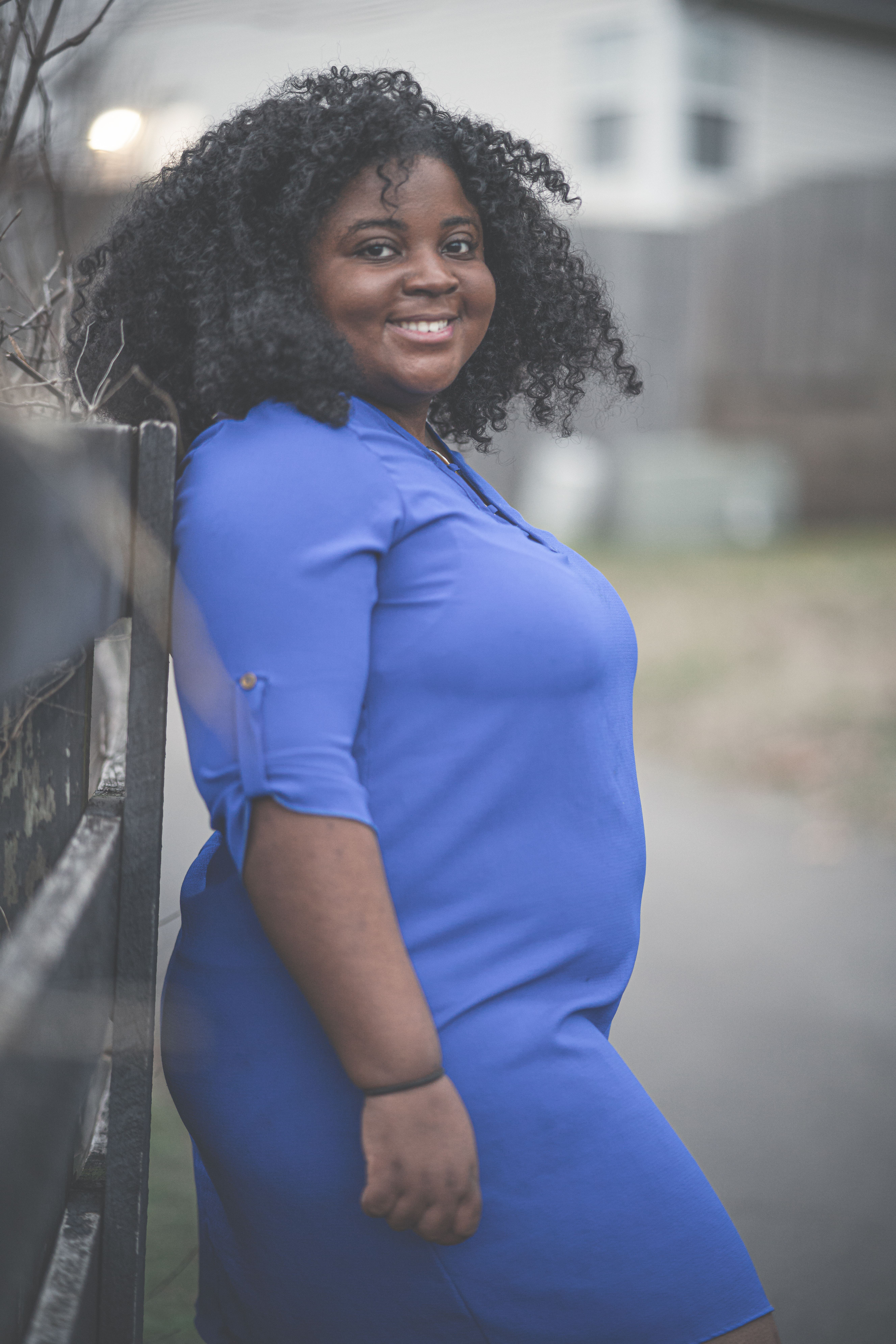 Smiling woman in a blue dress leaning against a fence, captured outdoors in daylight.