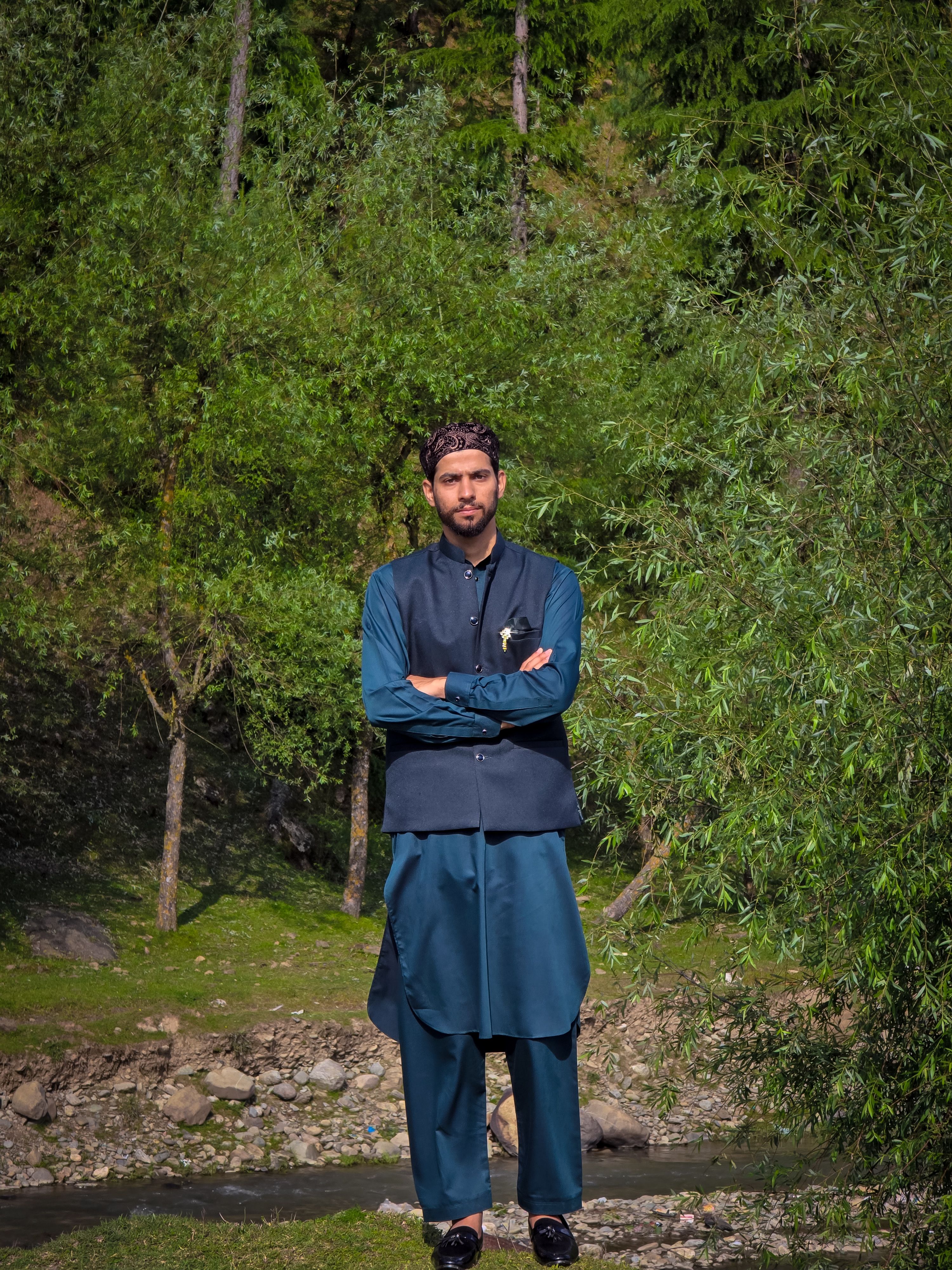 A man stands confidently in traditional attire amidst lush greenery, creating a serene outdoor scene.