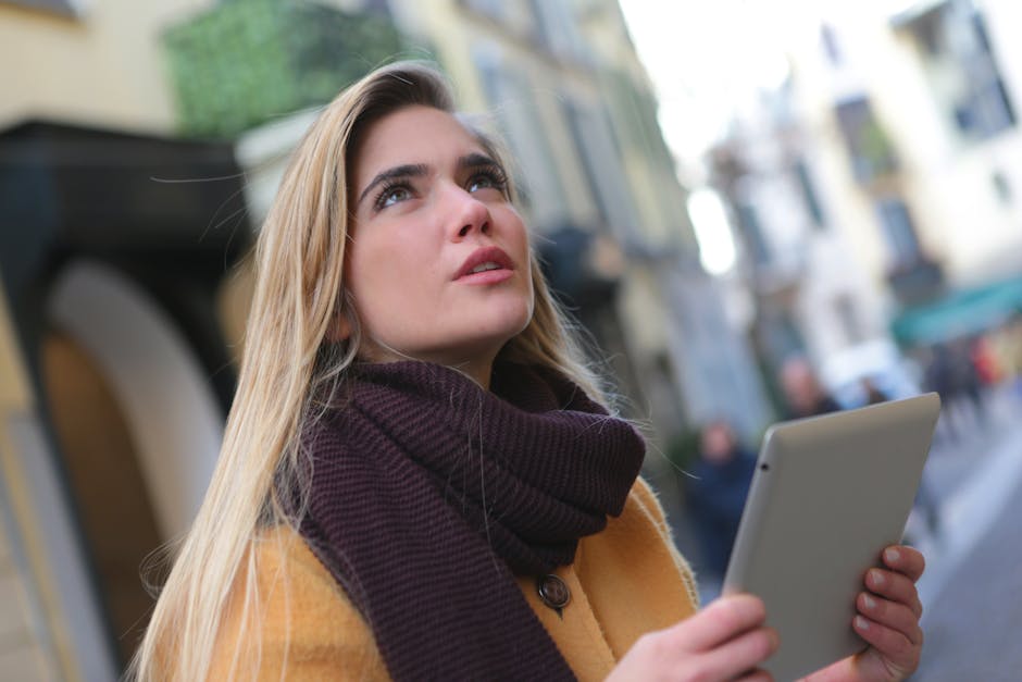 A young woman with a tablet looks up thoughtfully in a vibrant city street.
