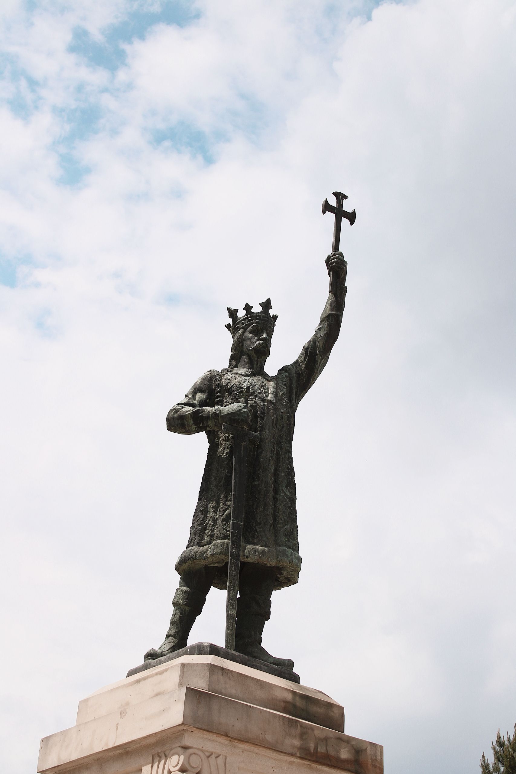 A towering statue of a monarch holding a cross dominates the scene, set against a cloudy sky.