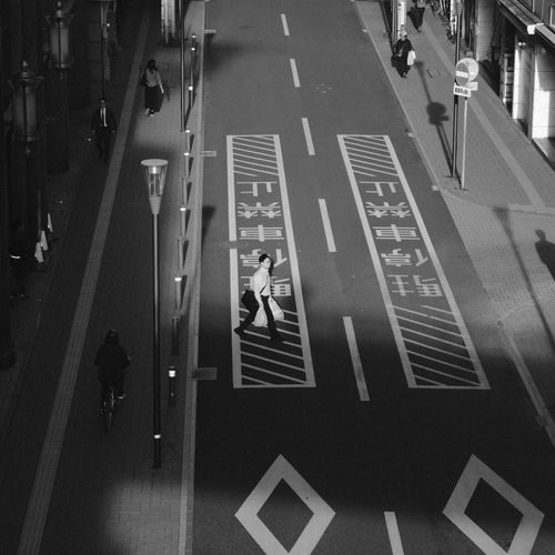 Free Black and white photo of a city street with pedestrians crossing, creating urban shadows. Stock Photo