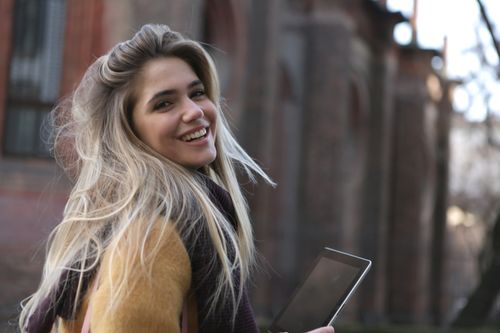 Free Happy young woman with blond hair smiling while holding a tablet outdoors. Stock Photo
