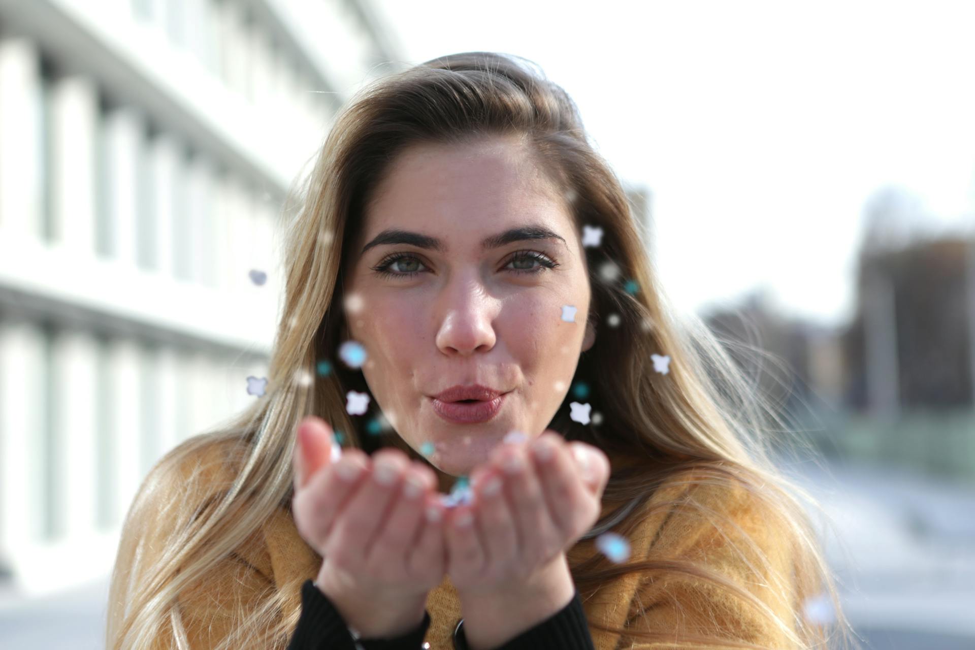 A young woman with blond hair blows confetti joyfully outdoors, capturing a playful moment.