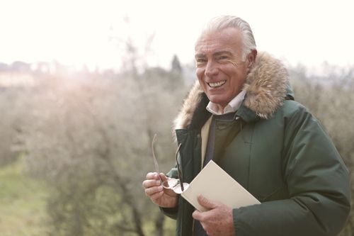 Free Senior man outdoors smiling warmly while holding a book and glasses. Stock Photo