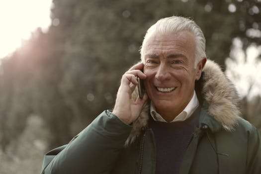 A smiling senior man in a fur-lined coat talks on his smartphone outdoors, embracing modern technology with joy.