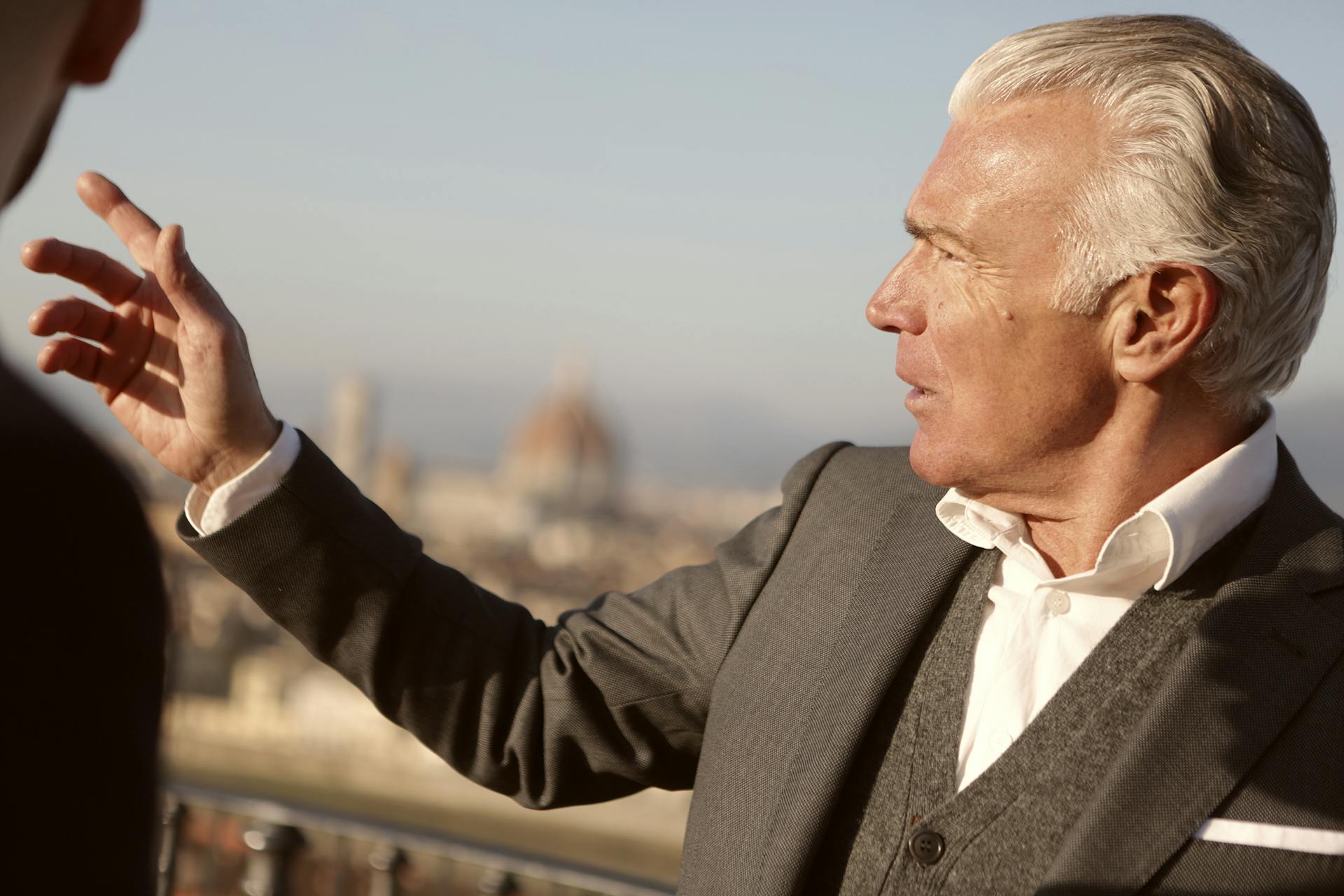 A senior businessman in formal attire gestures with Florence cityscape in the background.