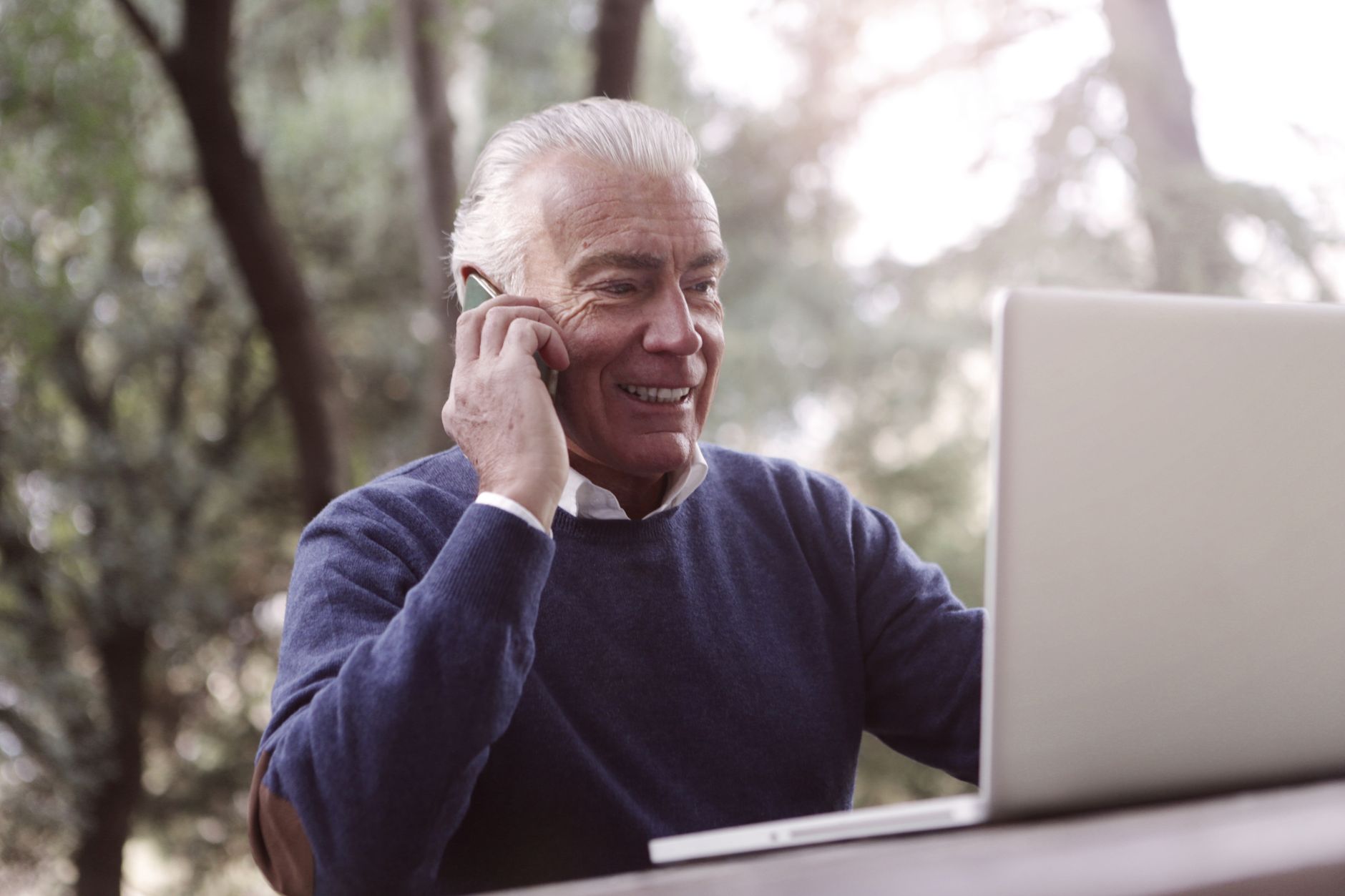 Elderly man smiling while on phone call with laptop outdoors.