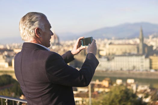 Elderly man captures city skyline with smartphone, enjoying a sunny day outdoors.