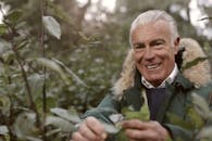 一位穿着绿色大衣的老人微笑着在秋季森林里探索，享受户外活动