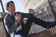 一名穿着牛仔夹克的男子坐在户外，微笑着在阳光下享用苹果