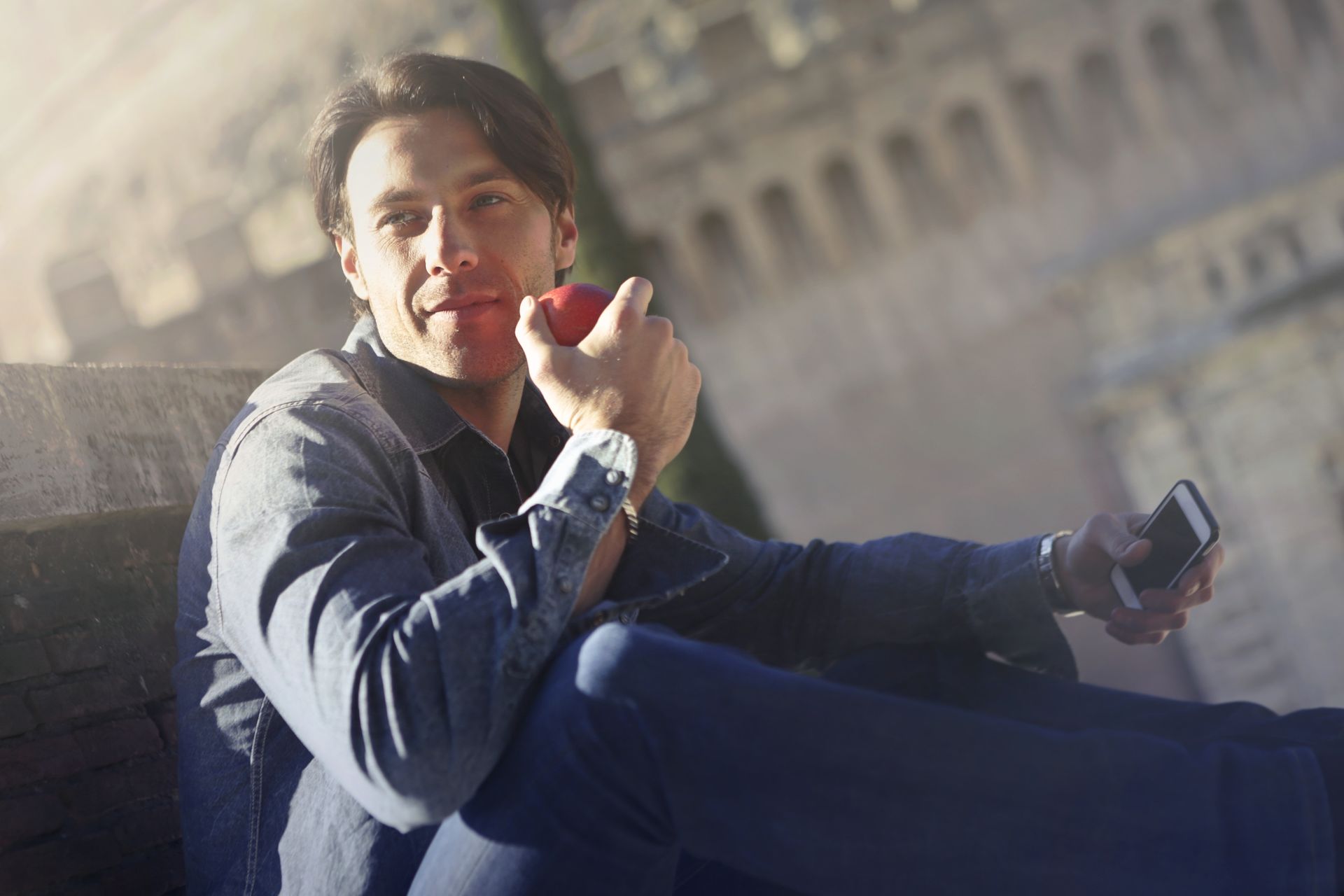 A young man enjoys a fresh apple and smartphone outdoors on a sunny day, embracing relaxation.