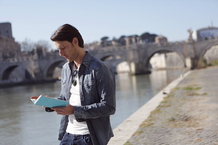 Young Man In Denim Jacket Is Reading A Book Near River Side