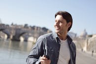 一个开朗的男人在阳光明媚的日子里，站在河边户外，背景是一座历史悠久的桥梁