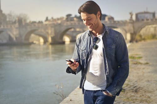 Man In Denim Jacket Holding A Smartphone Standing Near The River