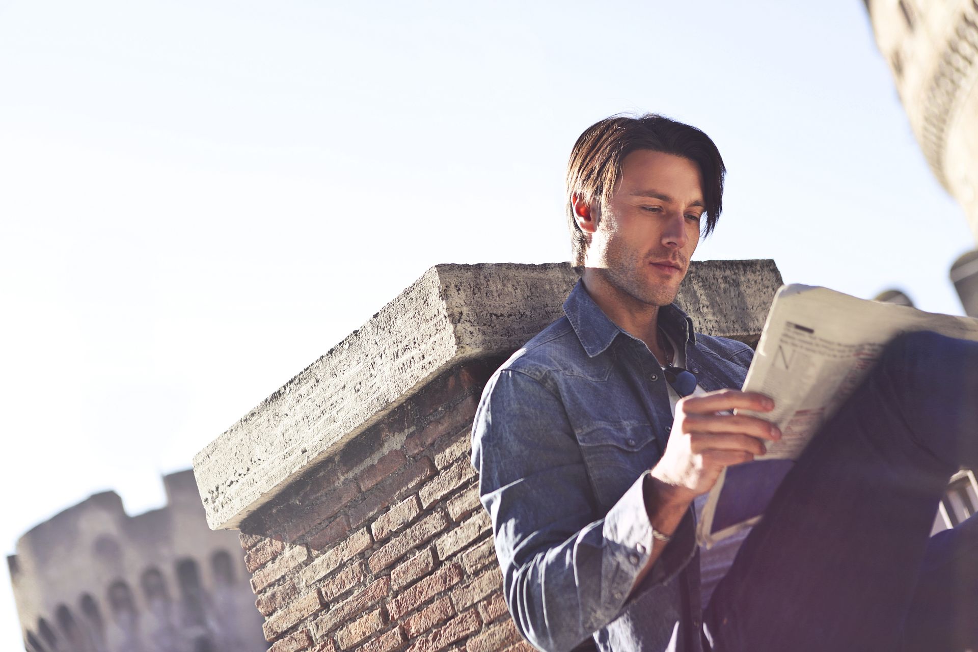 A man in a denim jacket reads a newspaper outdoors near a historic stone structure.