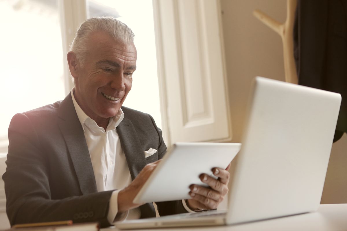 Smiling senior man multitasking with a laptop and tablet indoors at a desk